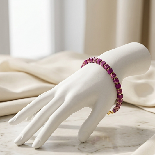 A white mannequin hand displays a beaded bracelet with alternating pink and purple beads and small gold accents, set against a soft cream fabric background.