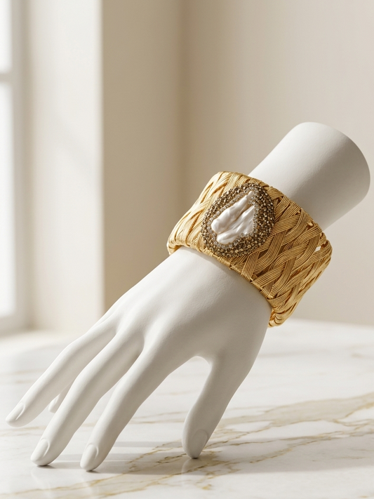 A wide gold cuff bracelet with a textured woven pattern and a central white shell inlay, displayed on a white mannequin hand over a light marble surface.