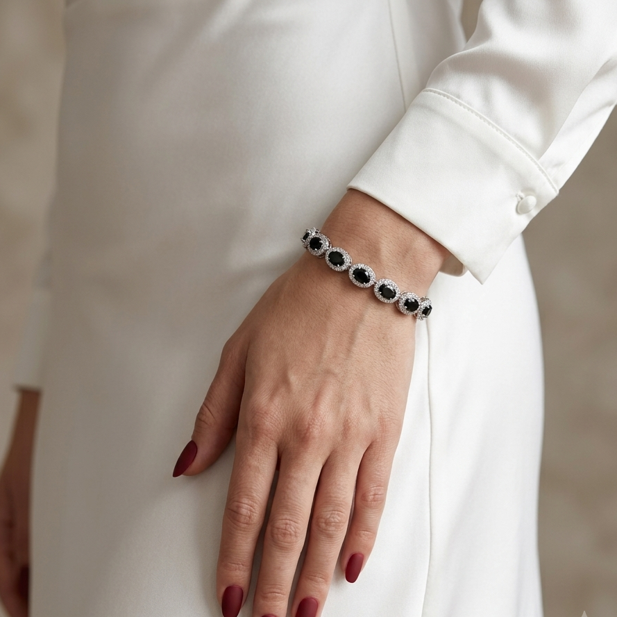 A close-up of a woman's hand wearing a delicate silver bracelet with oval black and white stones, styled with a white dress and dark red manicured nails.