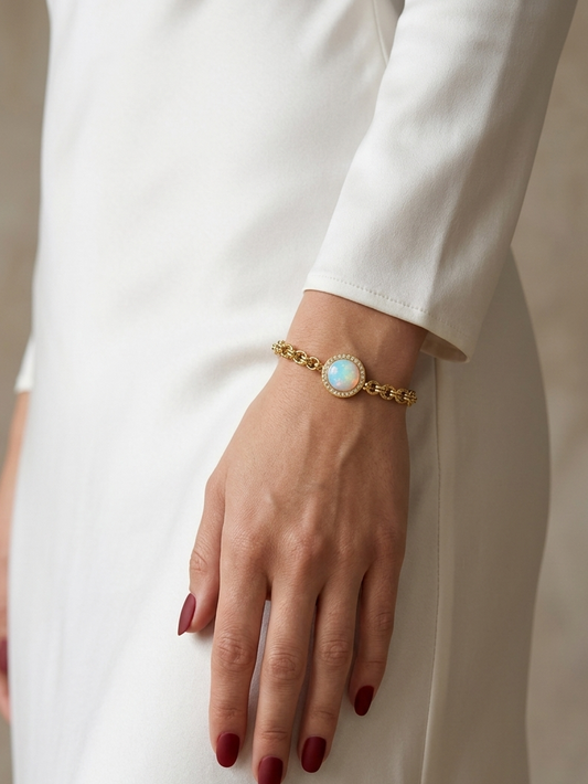 A gold chain bracelet with a round light blue cabochon stone, worn on a model's wrist. The model is wearing a white long-sleeved dress, and her hand has dark red nail polish.