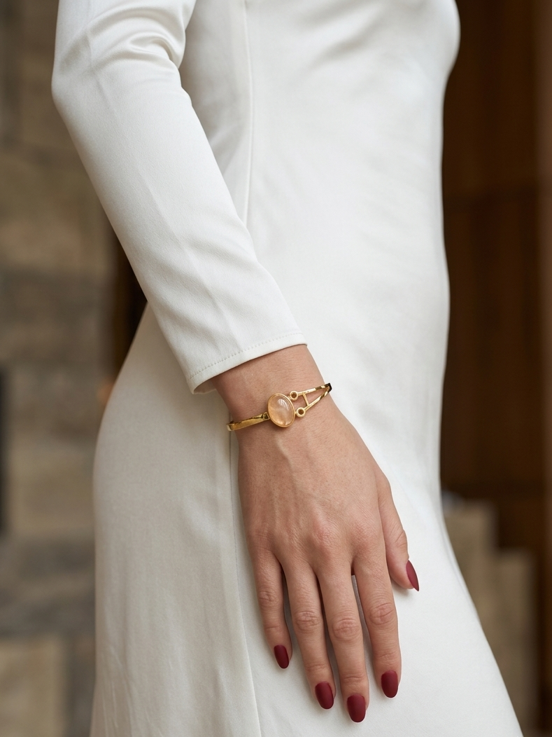 A close-up of a person wearing a delicate gold bangle bracelet with a circular beige stone centerpiece, styled with a long-sleeve white dress.