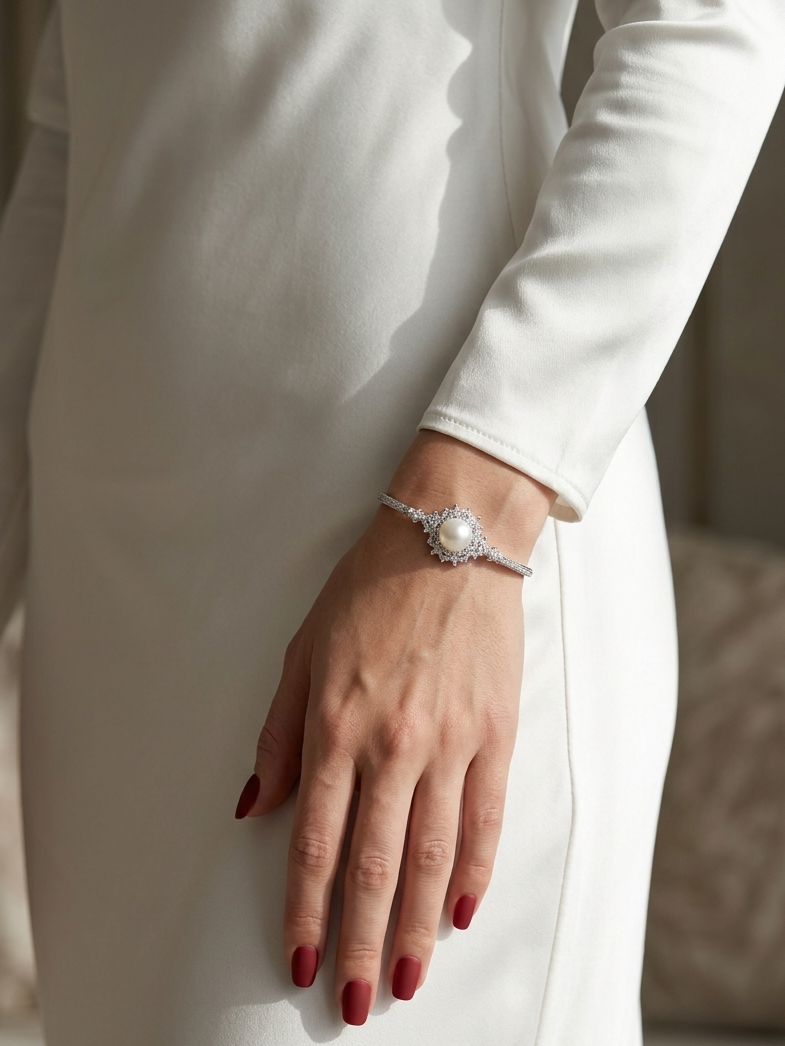 A close-up of a model's hand wearing a delicate silver bracelet with a central white pearl and surrounding clear stones, styled with a white long-sleeve dress.