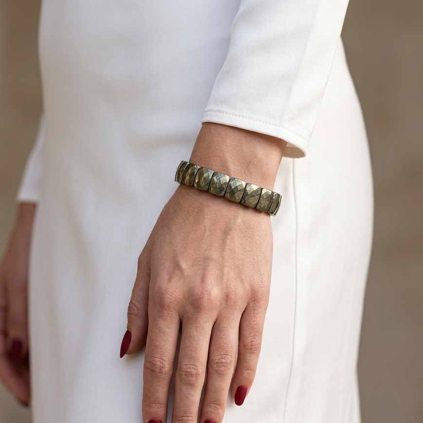 A close-up of a person wearing a chunky metallic cuff bracelet with a textured, hammered finish. The person is wearing a white long-sleeve dress, and the background is a soft, neutral beige wall.