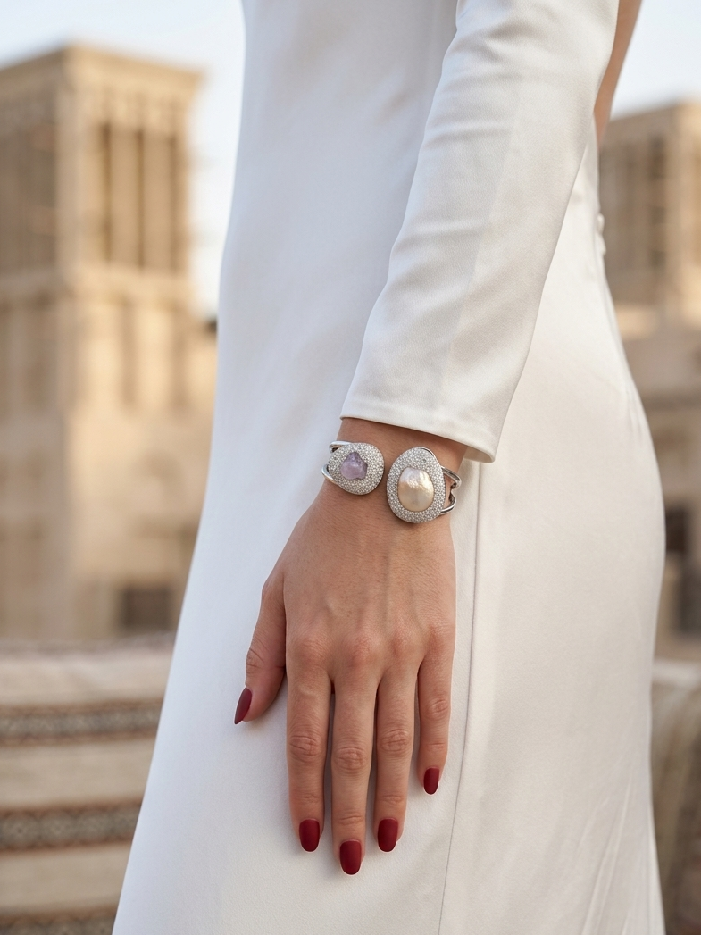 A close-up of a woman's hand with red manicured nails resting on her white dress, wearing a silver cuff bracelet with two large oval pearl-like centers and pinkish-purple inlay details. The background is a softly blurred outdoor architectural setting.