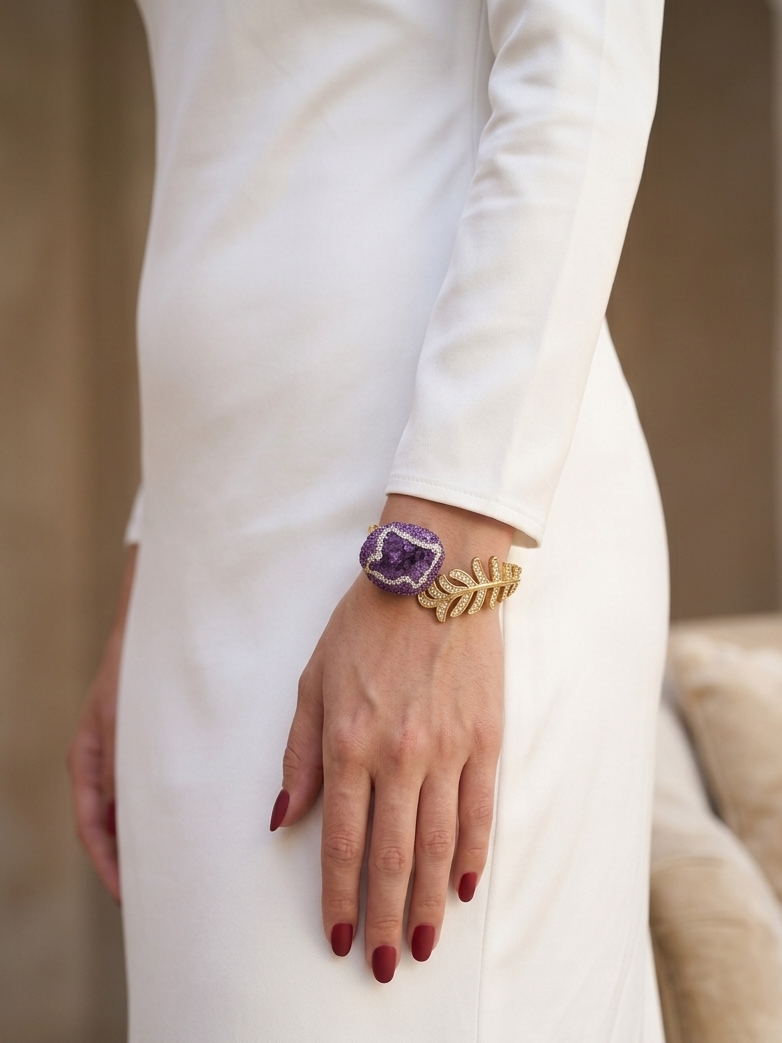 A close-up of a person wearing a white long-sleeve dress, showcasing a gold leaf bracelet with a purple amethyst crystal cabochon on the wrist.