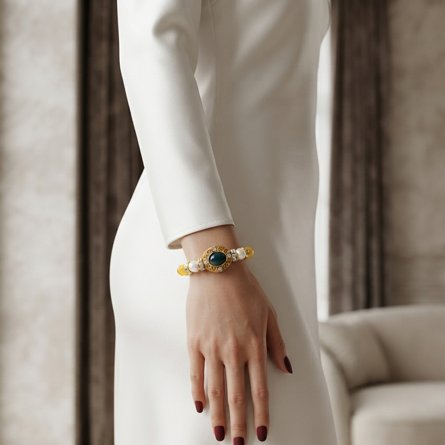 A woman in a white long-sleeve dress wears a gold-toned beaded bracelet with a central green oval stone and white pearl-like beads, photographed indoors near a window and sofa.