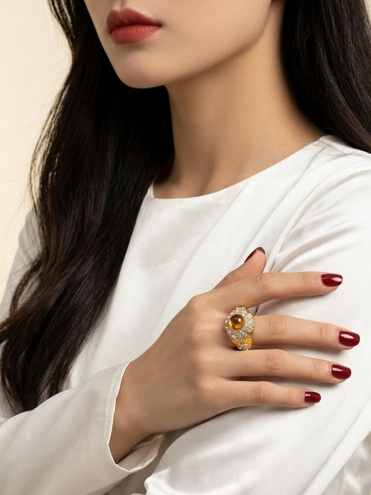 A close-up of a woman wearing a white satin long-sleeve top, showcasing a gold ring with a central amber-brown stone and small clear stones on her hand.