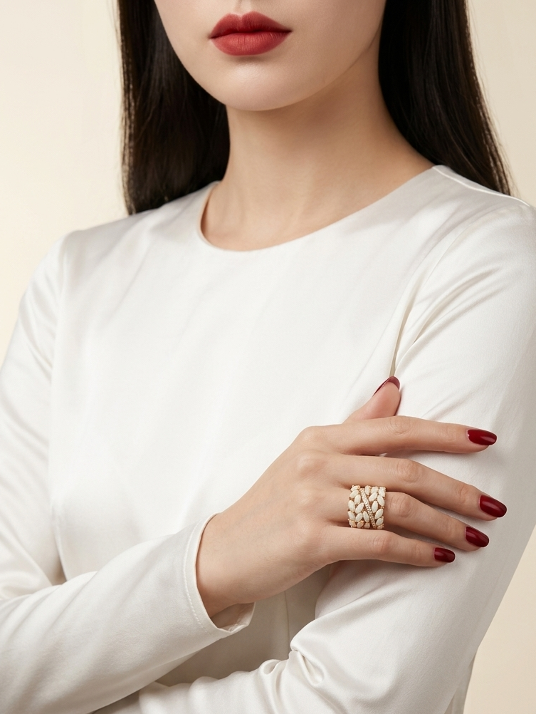 A close-up of a woman wearing a white long-sleeve top and a textured gold ring on her finger, set against a soft beige background.