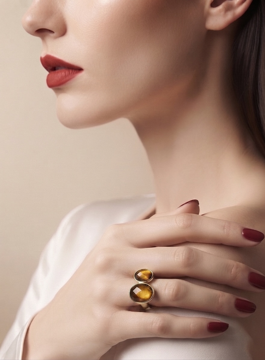 A close-up lifestyle shot of a model wearing a double-stone statement ring. The ring features two polished oval yellow-gold cabochons in a bezel setting with a dark metallic border, worn on her ring finger.