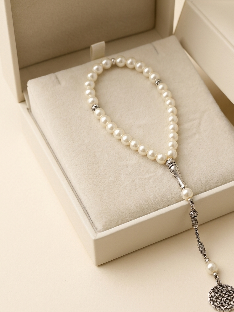 A pearl beaded necklace with silver-toned spacer beads and a dangling silver-toned tassel charm, displayed inside an open cream-colored jewelry box.