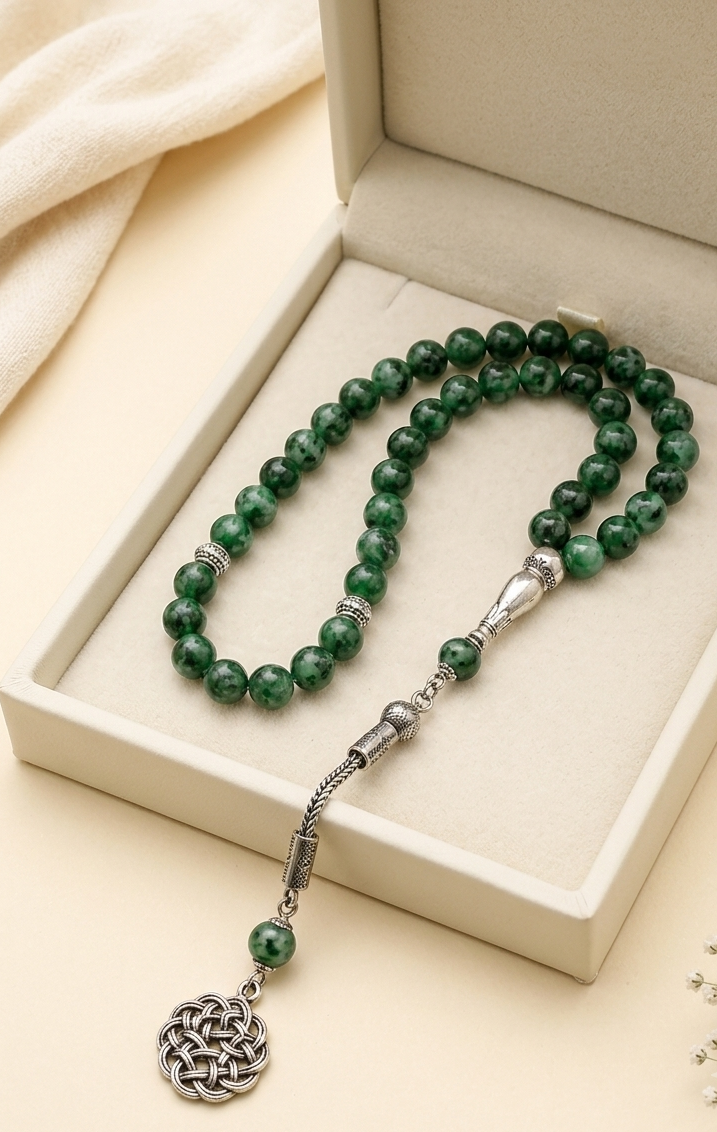 A beaded necklace with dark green gemstone-like beads and silver-toned metal accents, displayed inside an open cream-colored jewelry box. The necklace includes a dangling silver-toned Celtic knot charm.