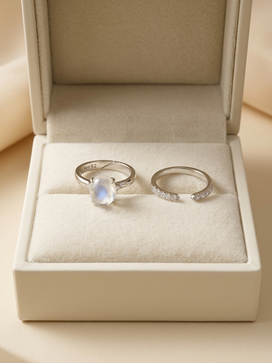Two silver rings displayed in an open cream-colored jewelry box. One ring features a round iridescent moonstone, and the other is a delicate open band set with small clear stones.