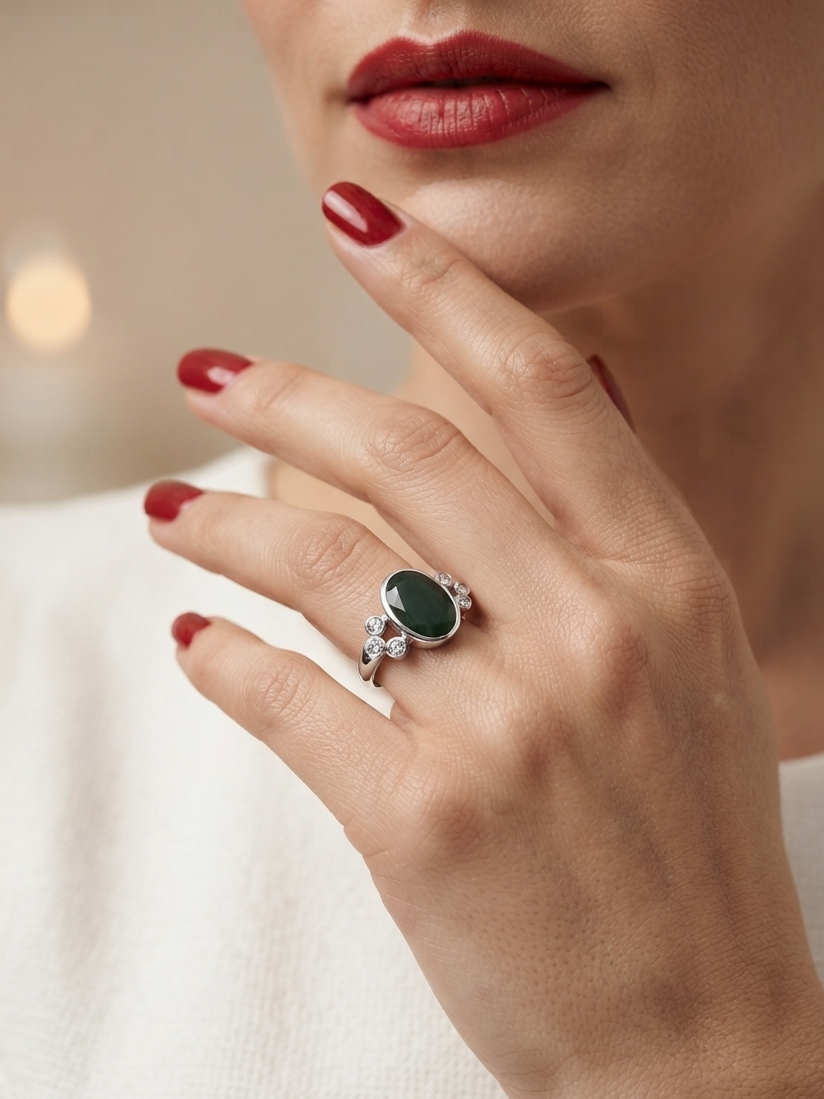 A close-up lifestyle shot of a model's hand wearing a silver ring with a dark green oval cabochon and small clear accent stones, set against a soft, warm blurred background.