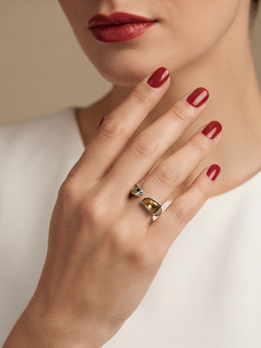 A close-up lifestyle shot of a model's hand wearing a silver ring with a large, polished yellow gemstone. The model has red nail polish and is wearing a white top against a soft beige background.