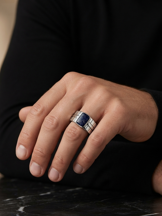 A close-up of a man's hand wearing a silver-toned ring with a rectangular dark blue inlay, resting on a dark marble surface.