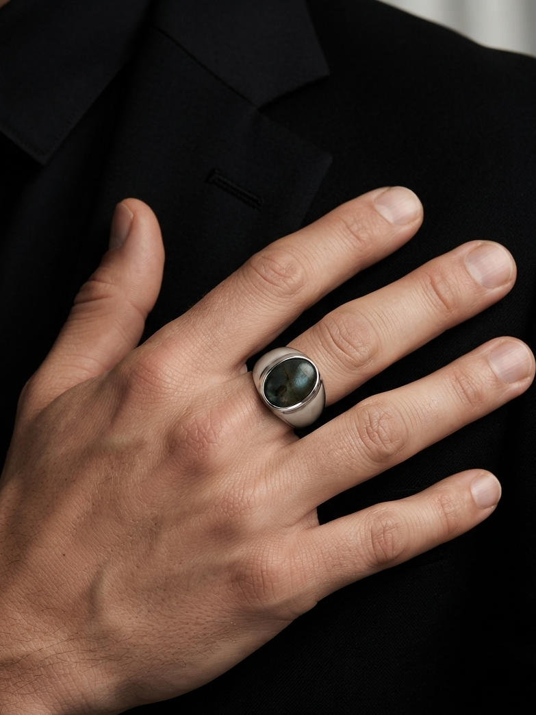 A silver-toned men's ring with a dark oval cabochon stone, shown worn on a man's ring finger over a black suit jacket.