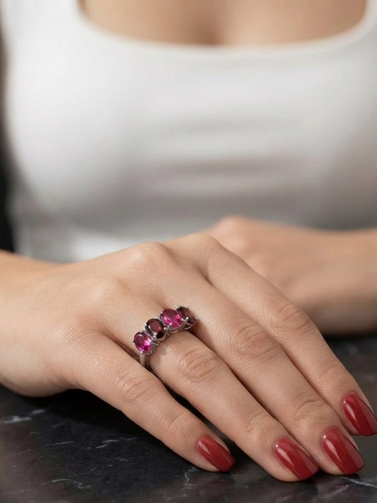 A close-up of a woman's hand resting on a dark marble surface, wearing a silver ring set with three oval pink gemstones. The person is wearing a white top, and the background is a soft, blurred neutral tone.