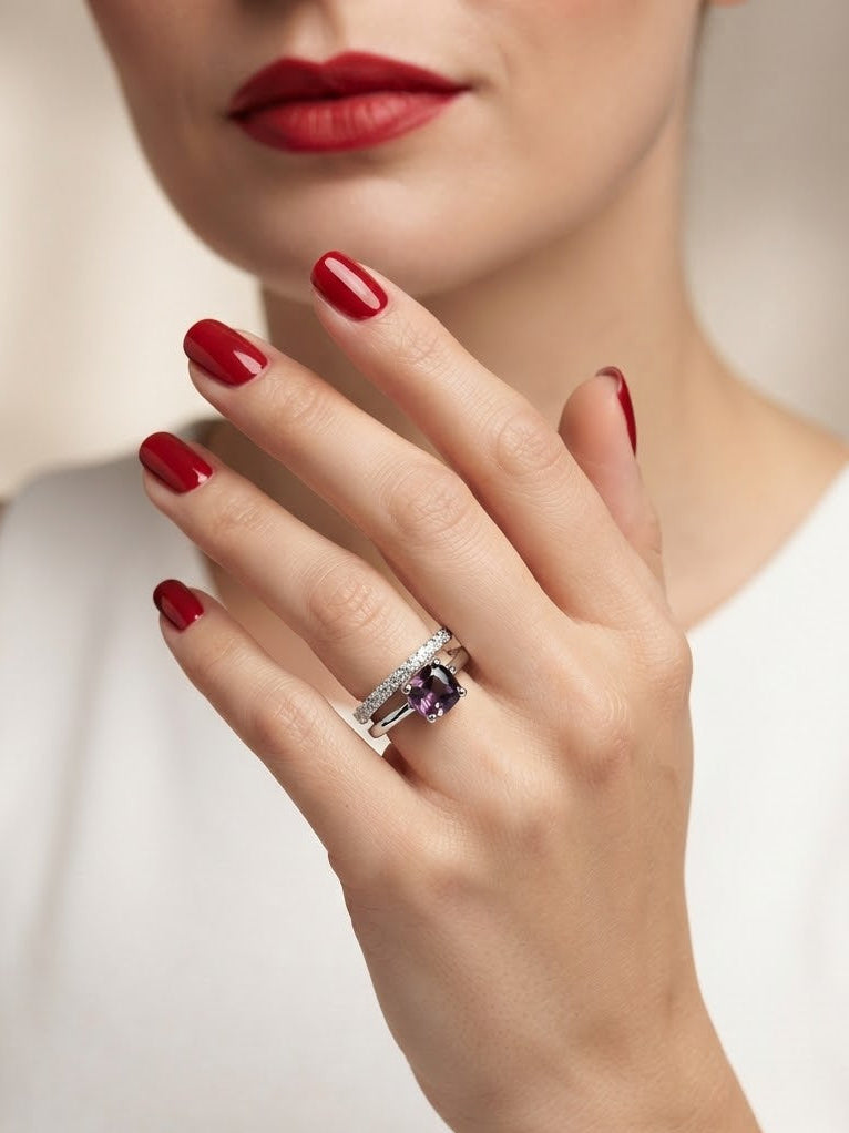 A close-up of a model's hand wearing a silver-toned ring with a prominent oval purple gemstone, set against a soft beige background and white top.