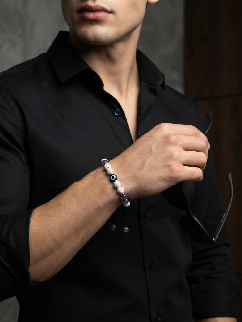 A man wearing a black button-up shirt and holding sunglasses while wearing a beaded bracelet on his wrist. The bracelet features white pearl-like beads, a blue evil eye bead, and purple beads.