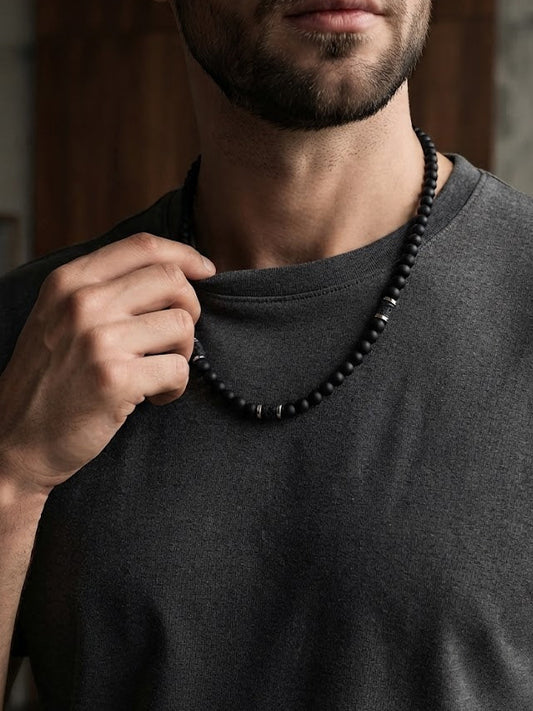 A close-up of a man wearing a dark charcoal gray t-shirt and a black beaded necklace, with his hand adjusting the collar of his shirt.