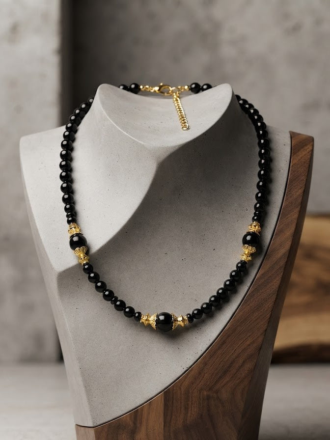 A black beaded necklace with gold-toned spacer beads and a small dangling charm, displayed on a gray jewelry bust with a wooden base against a softly blurred indoor background.