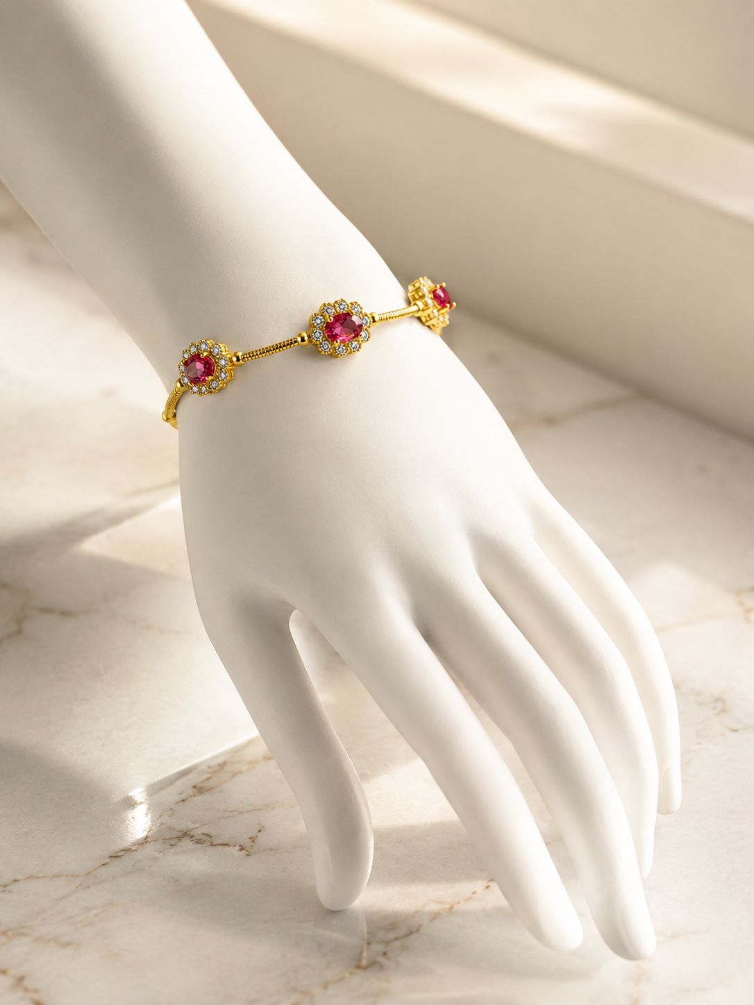 A gold-toned bangle bracelet with three red gemstone floral motifs, displayed on a white mannequin hand over a light marble surface.