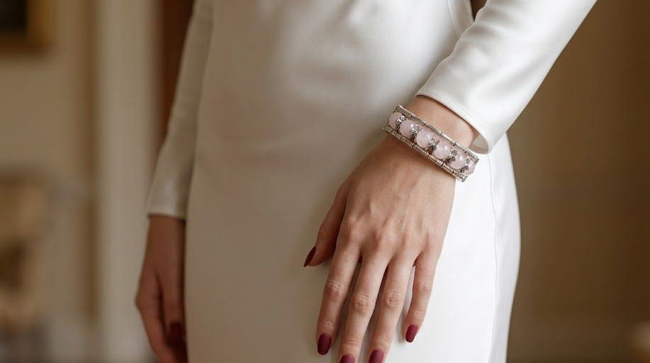 A close-up of a person wearing a white long-sleeve dress and a wide silver cuff bracelet with pink gemstones, with the hand resting on the hip.