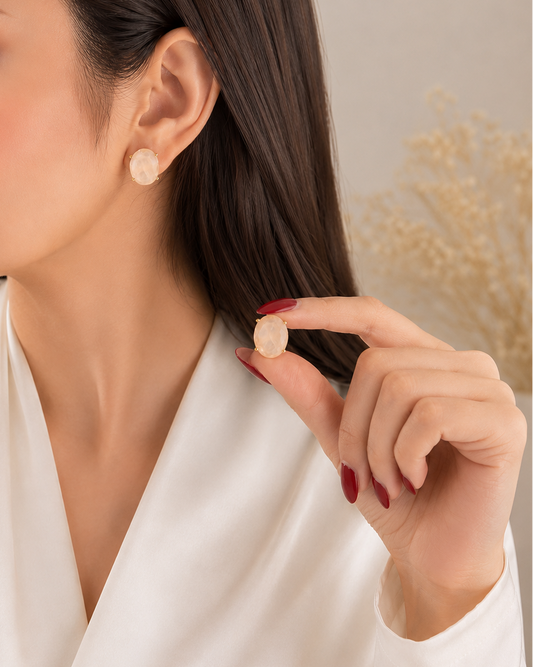 A close-up lifestyle shot of a woman wearing a small, round, textured cream-colored stud earring, holding another one near her ear. She is wearing a white satin robe against a soft beige background with dried floral elements.