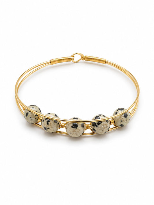 A gold wire bangle bracelet featuring five round, speckled Dalmatian stone beads spaced along the front, set against a plain white background.