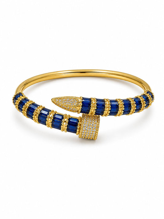 A gold bangle bracelet featuring deep blue rectangular gemstones and pavé-set clear stones, photographed on a clean white background.