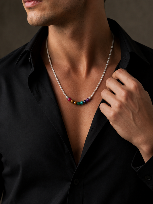 A close-up of a man wearing a silver chain necklace with a small horizontal row of colorful round beads at the center, paired with an open black button-down shirt.