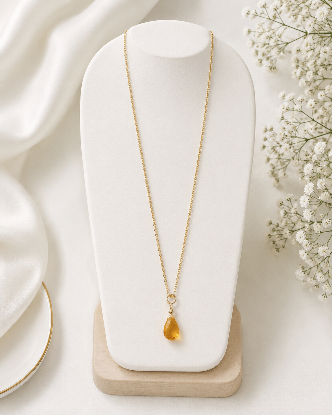 Gold necklace with a yellow pendant on a white stand with white flowers in the background