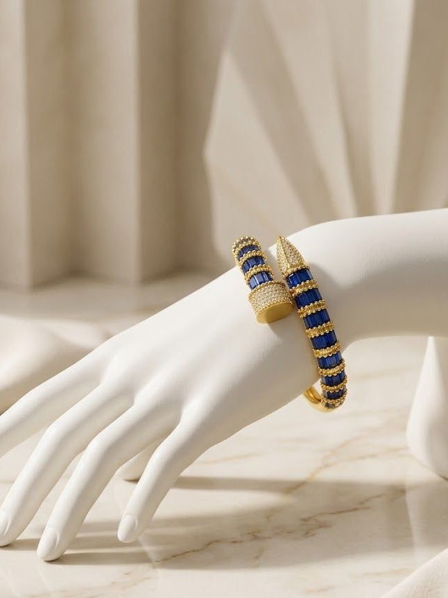 A gold-toned cuff bracelet with alternating deep blue and textured gold segments, displayed on a white mannequin hand against a soft beige draped background.