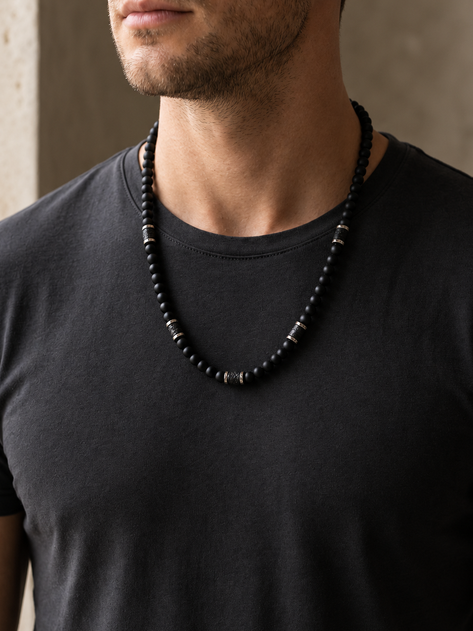 A close-up of a man wearing a black beaded necklace with silver-toned spacer beads over a dark gray t-shirt.