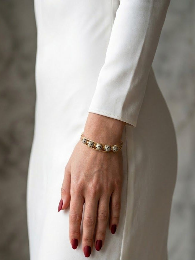 A close-up of a woman's hand wearing a delicate gold bracelet with small clear stones, styled with a white long-sleeve dress and dark red manicured nails.