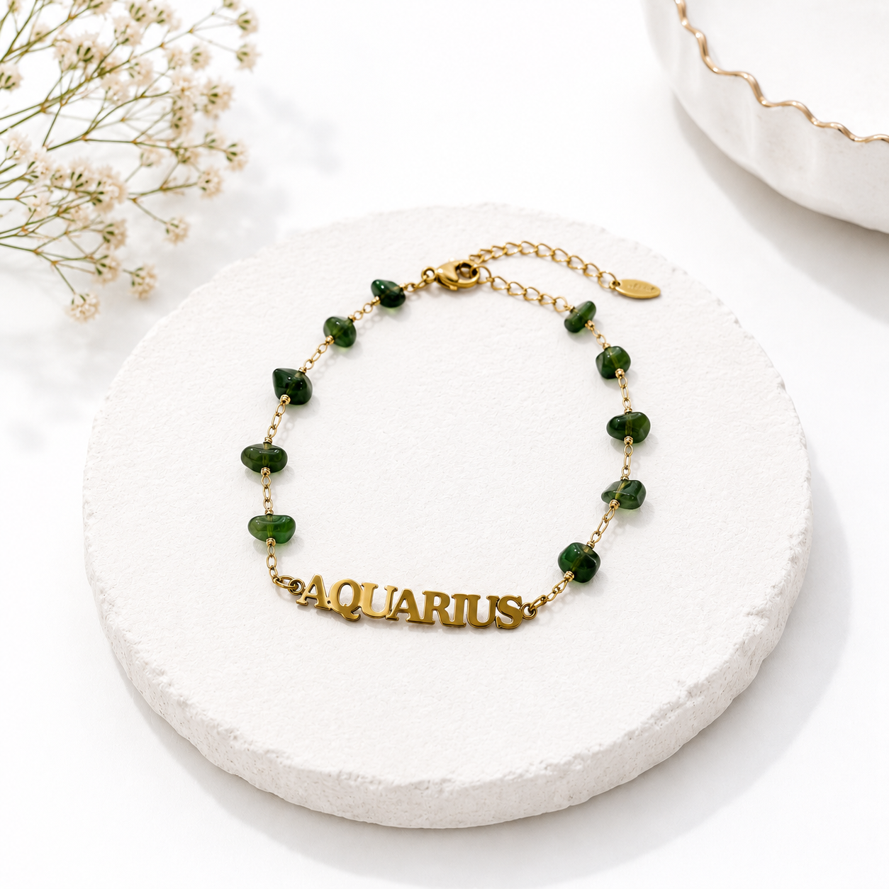 Gold bracelet with green beads labeled 'Aquarius' on a white surface with baby's breath flowers.