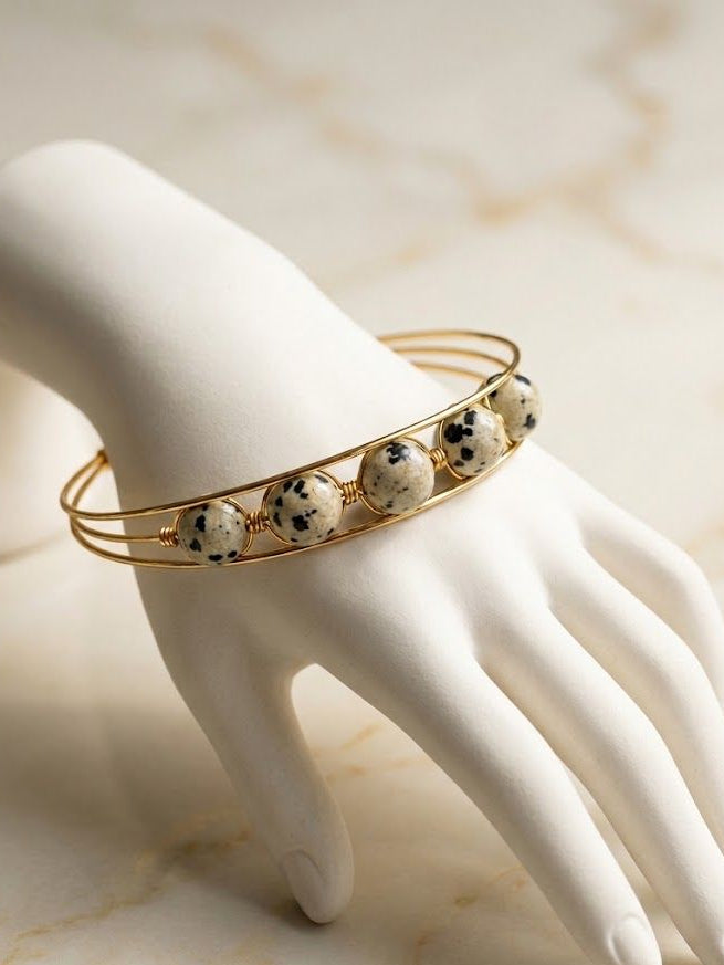 A gold-toned double-band cuff bracelet with five round speckled white beads, displayed on a white mannequin hand against a light marble background.
