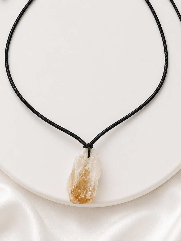 A black cord necklace with a raw, polished crystal pendant in warm beige and amber tones, displayed on a white circular stand.