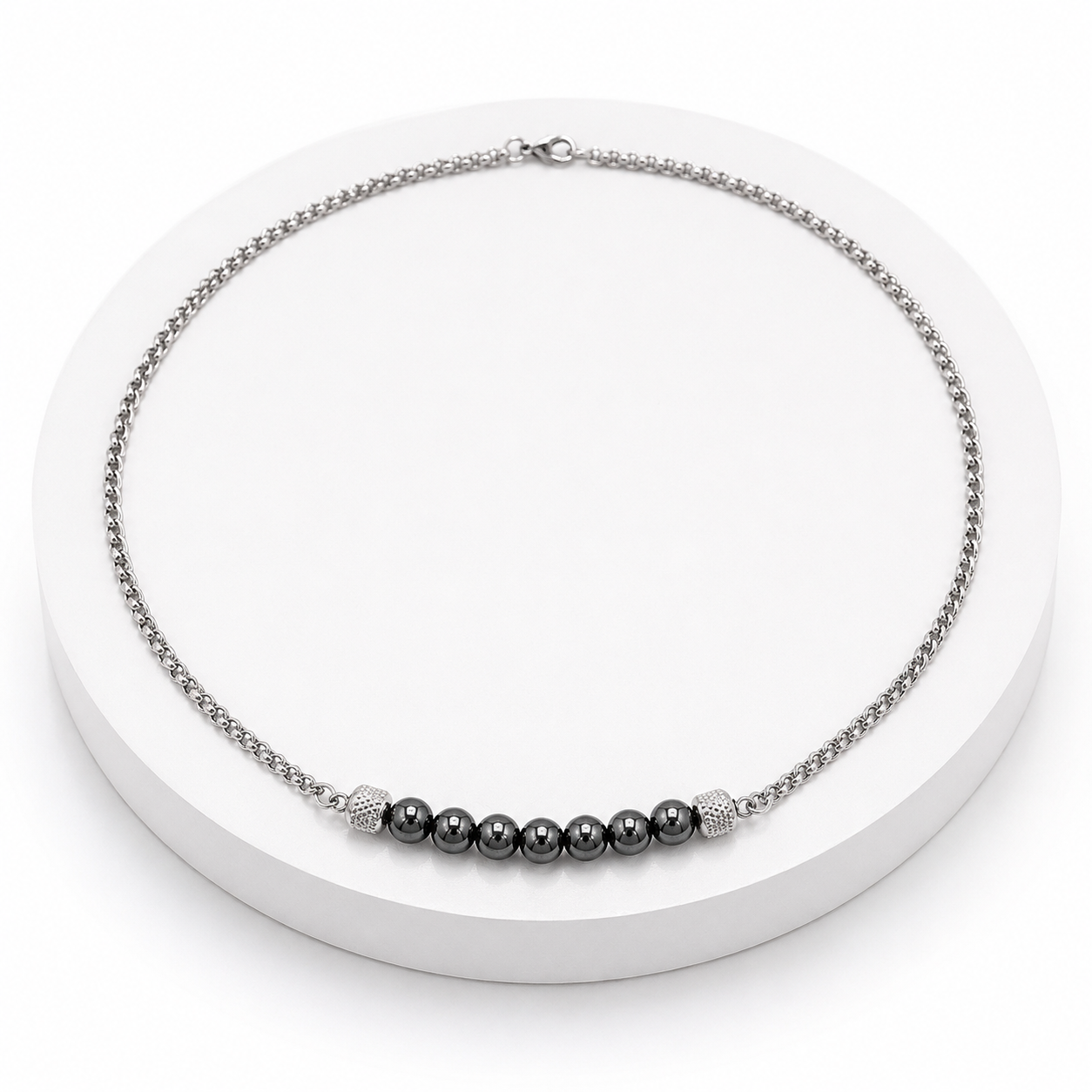 A silver-toned chain necklace with a central row of dark gray beads and textured silver end caps, displayed on a white circular pedestal.