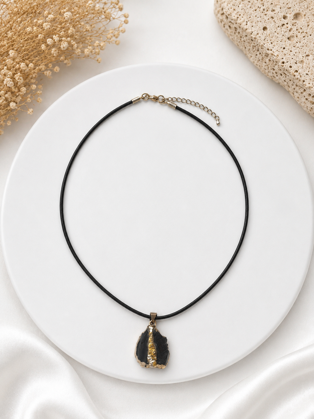 Necklace with a black cord and pendant on a white plate with beige flowers and fabric in the background