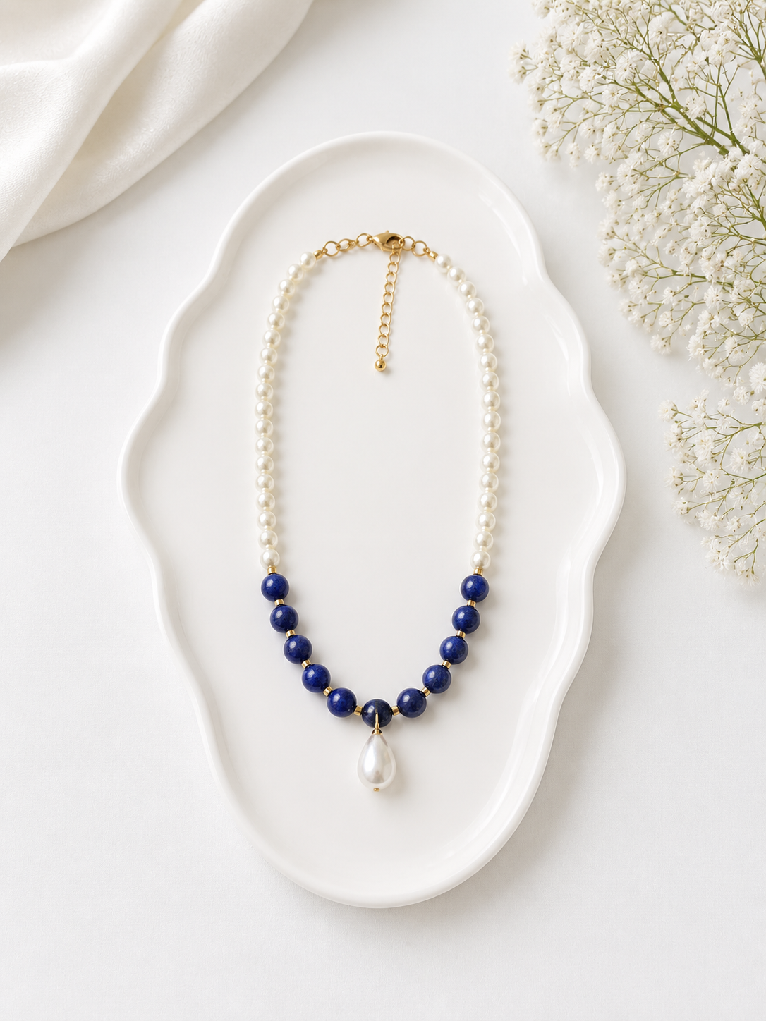 Necklace with blue beads and a pearl on a white dish with flowers in the background
