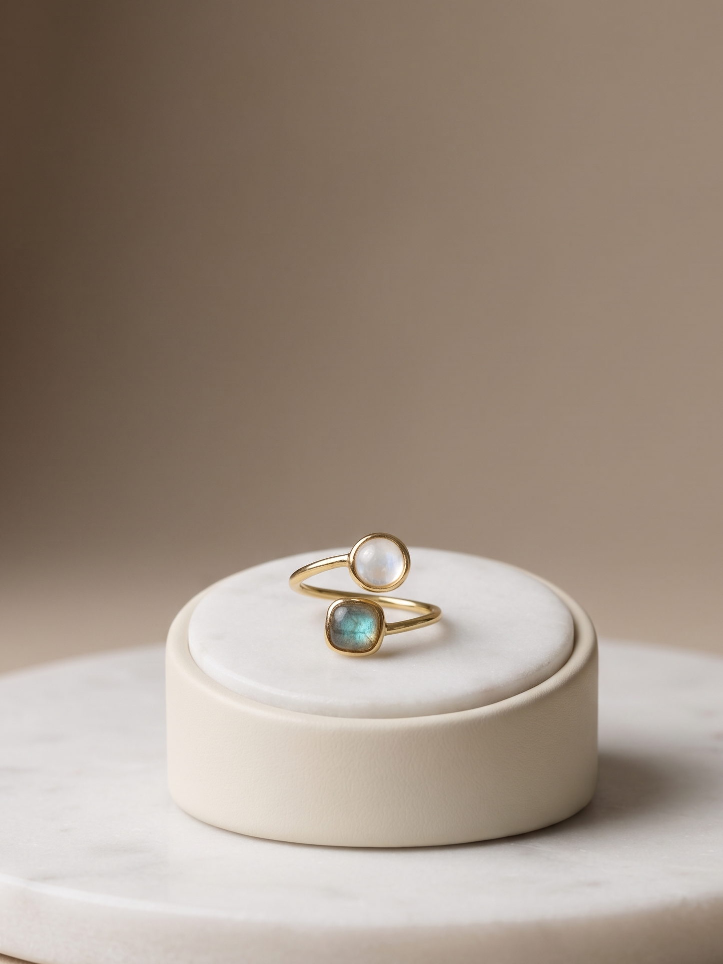 Two gold rings displayed on a small round cream pedestal. One ring has a smooth white pearl, and the other has a teardrop-shaped iridescent blue-green stone.
