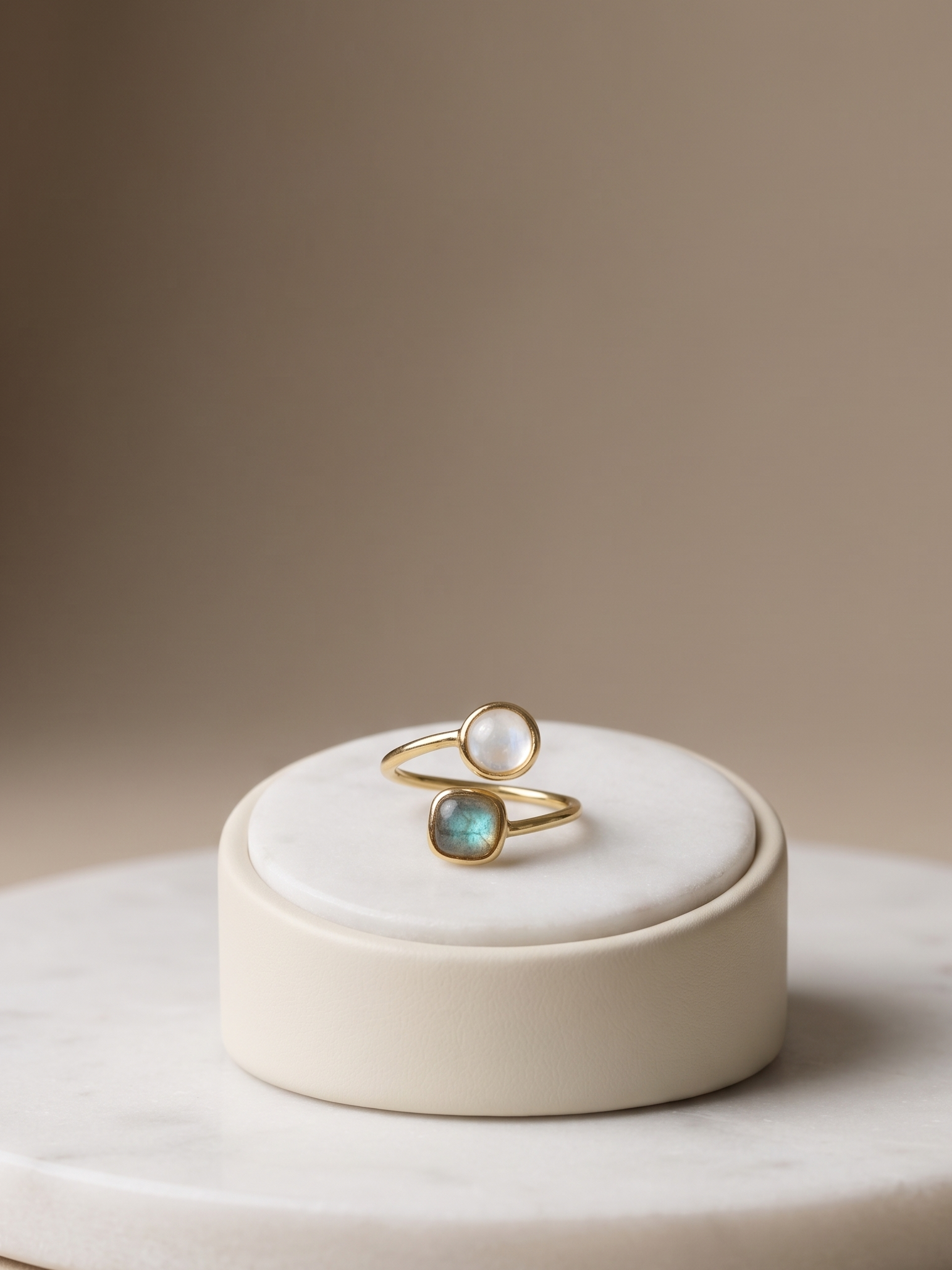 Two gold rings displayed on a small round cream pedestal. One ring has a smooth white pearl, and the other has a teardrop-shaped iridescent blue-green stone.