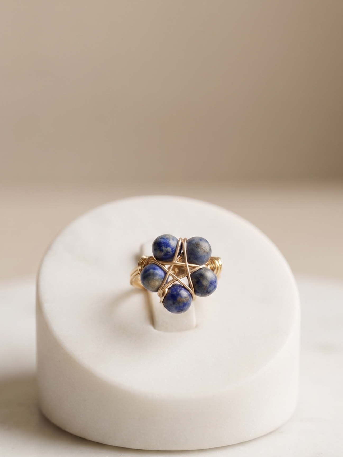 A gold ring with four dark blue gemstone clusters, displayed on a white ceramic ring stand over a light marble surface.