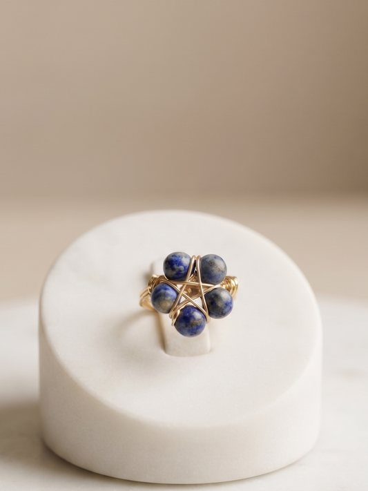 A gold ring with four dark blue gemstone clusters, displayed on a white ceramic ring stand over a light marble surface.