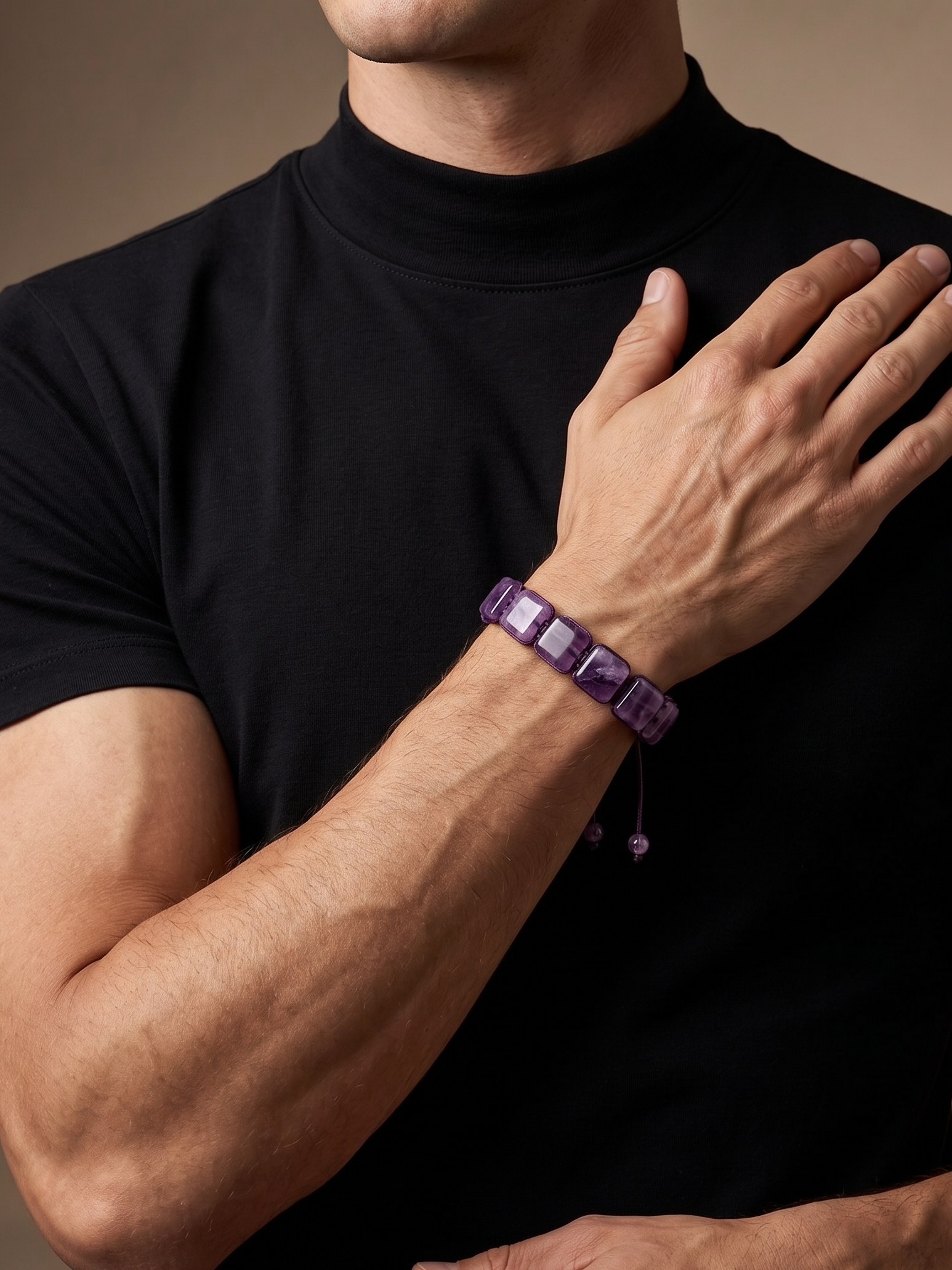 A close-up of a man wearing a black turtleneck shirt, showcasing a purple beaded bracelet on his wrist against a warm, neutral background.