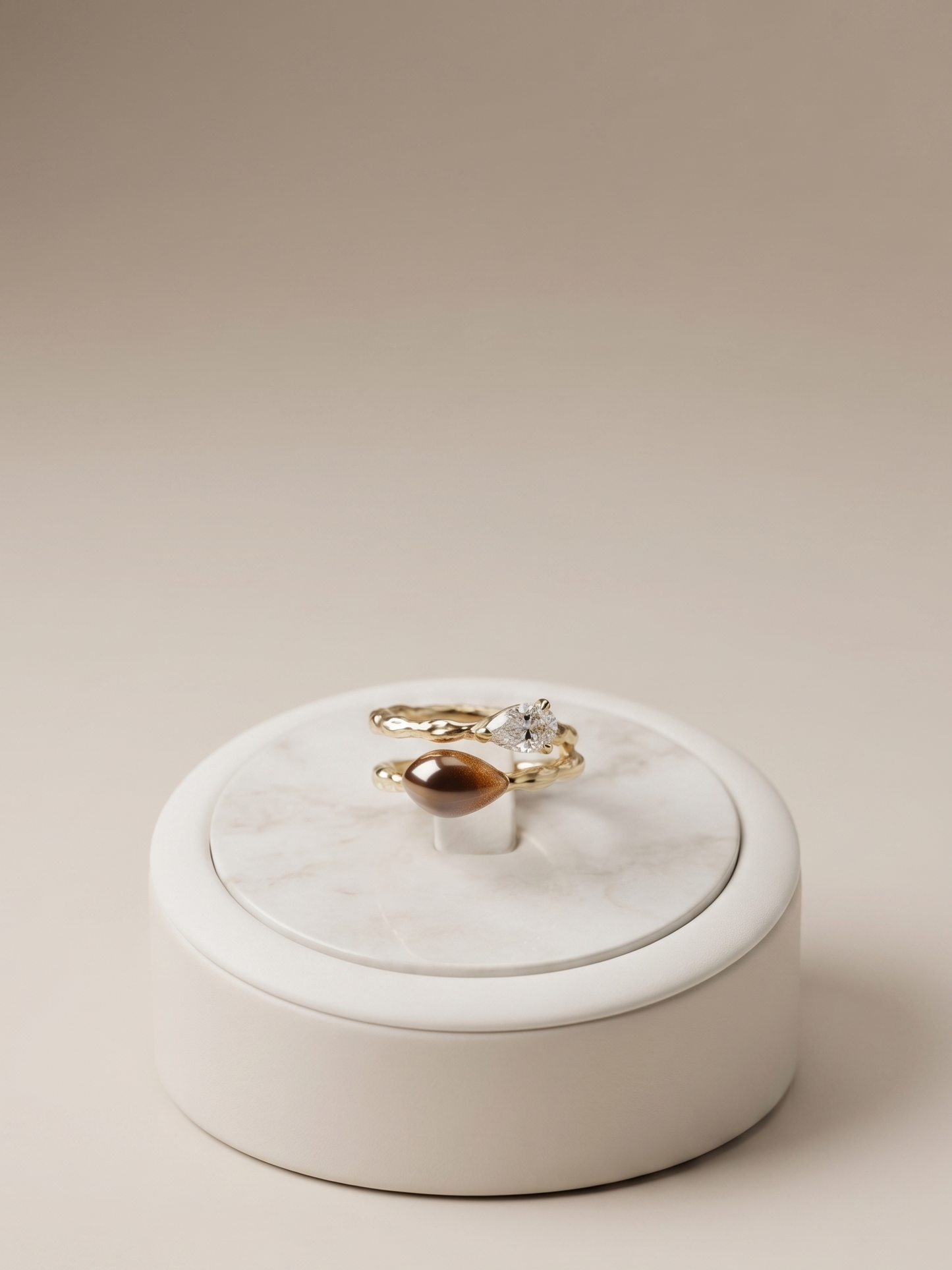 Two gold rings, one with a marquise-cut gemstone and one with a round brown gemstone, displayed on a white marble and cream ceramic ring box against a warm beige background.
