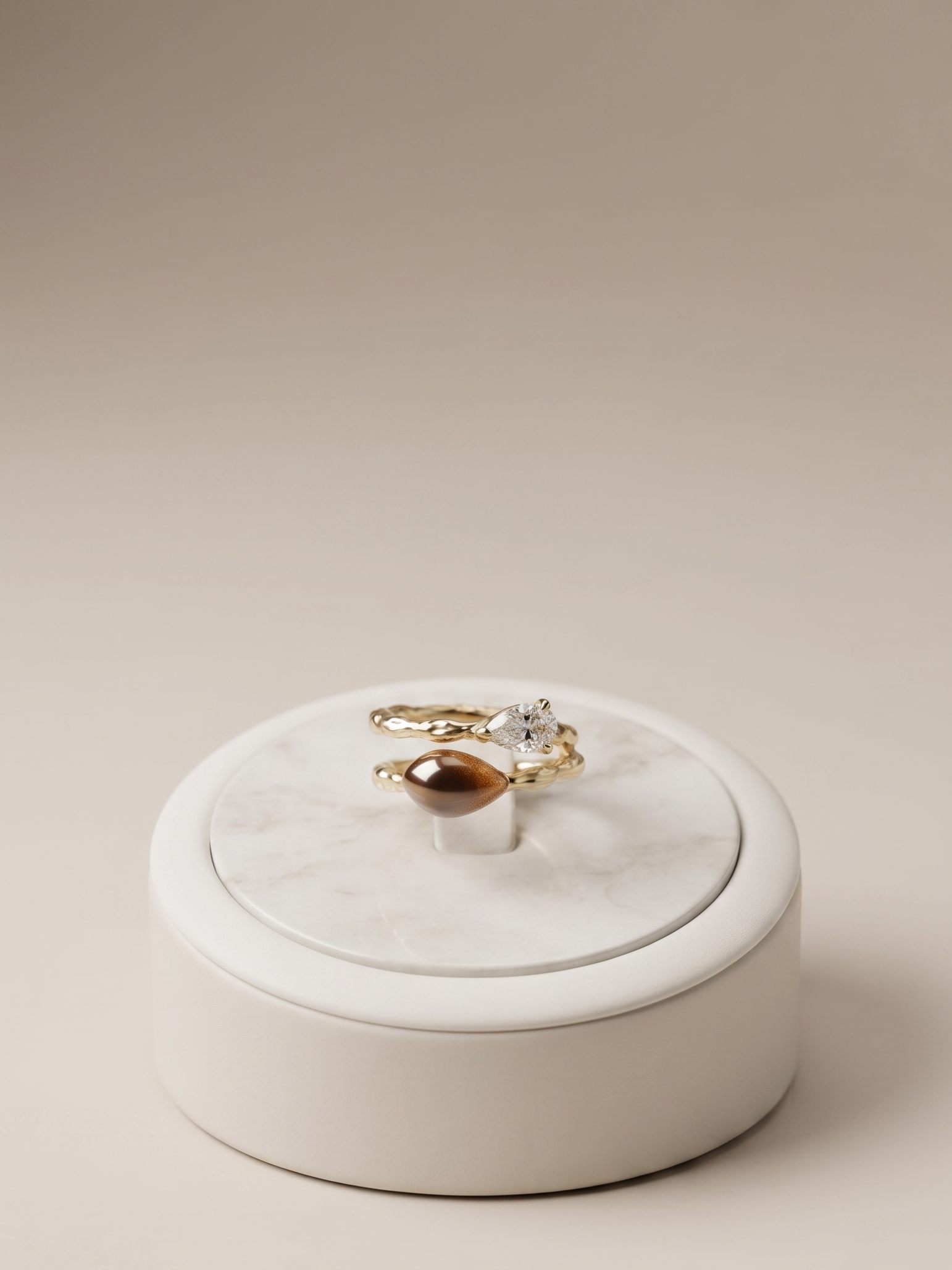 Two gold rings, one with a marquise-cut gemstone and one with a round brown gemstone, displayed on a white marble and cream ceramic ring box against a warm beige background.