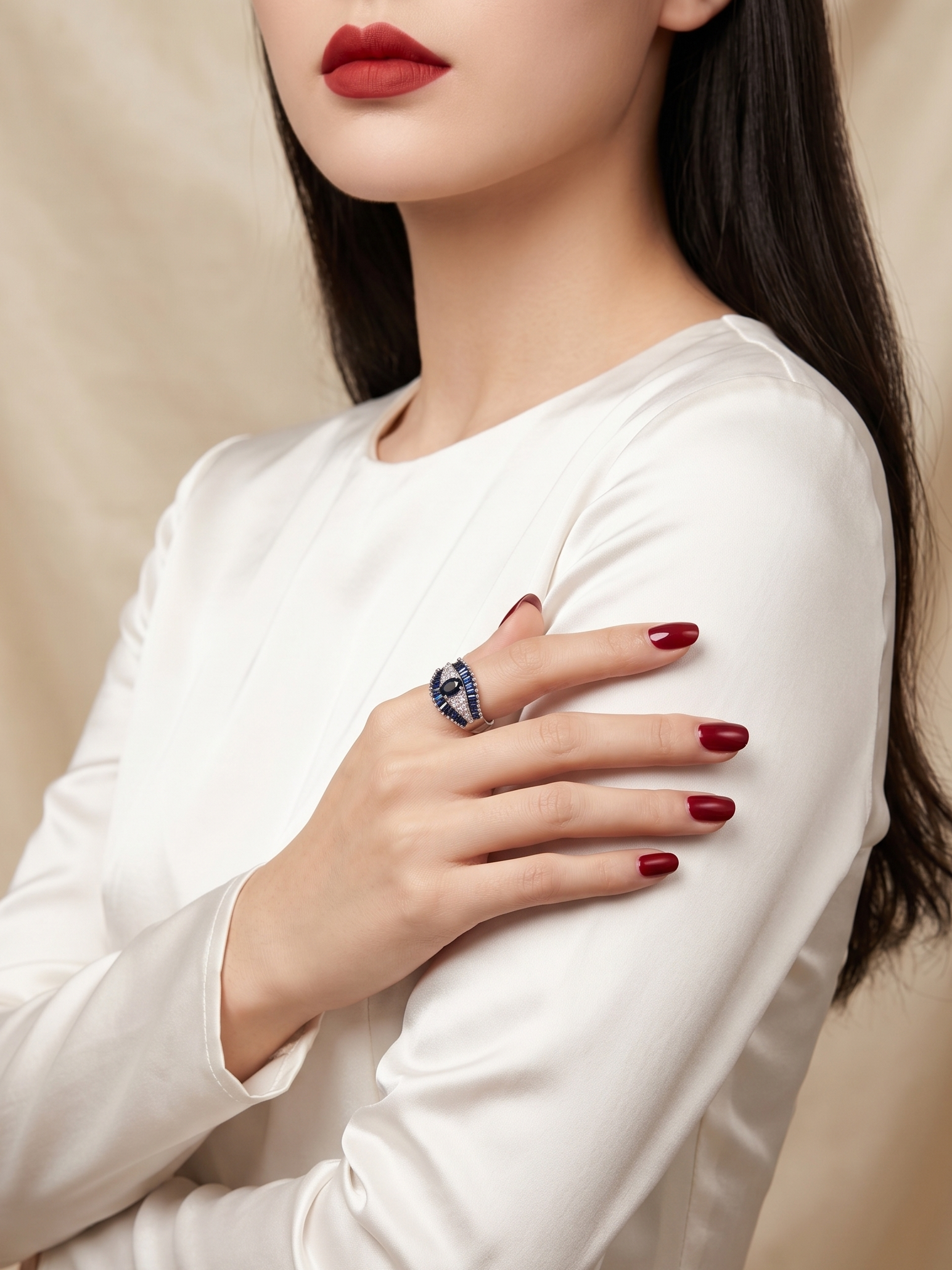 A close-up of a woman wearing a white long-sleeve top and a dark ring with a blue stone, posed against a soft beige fabric backdrop.