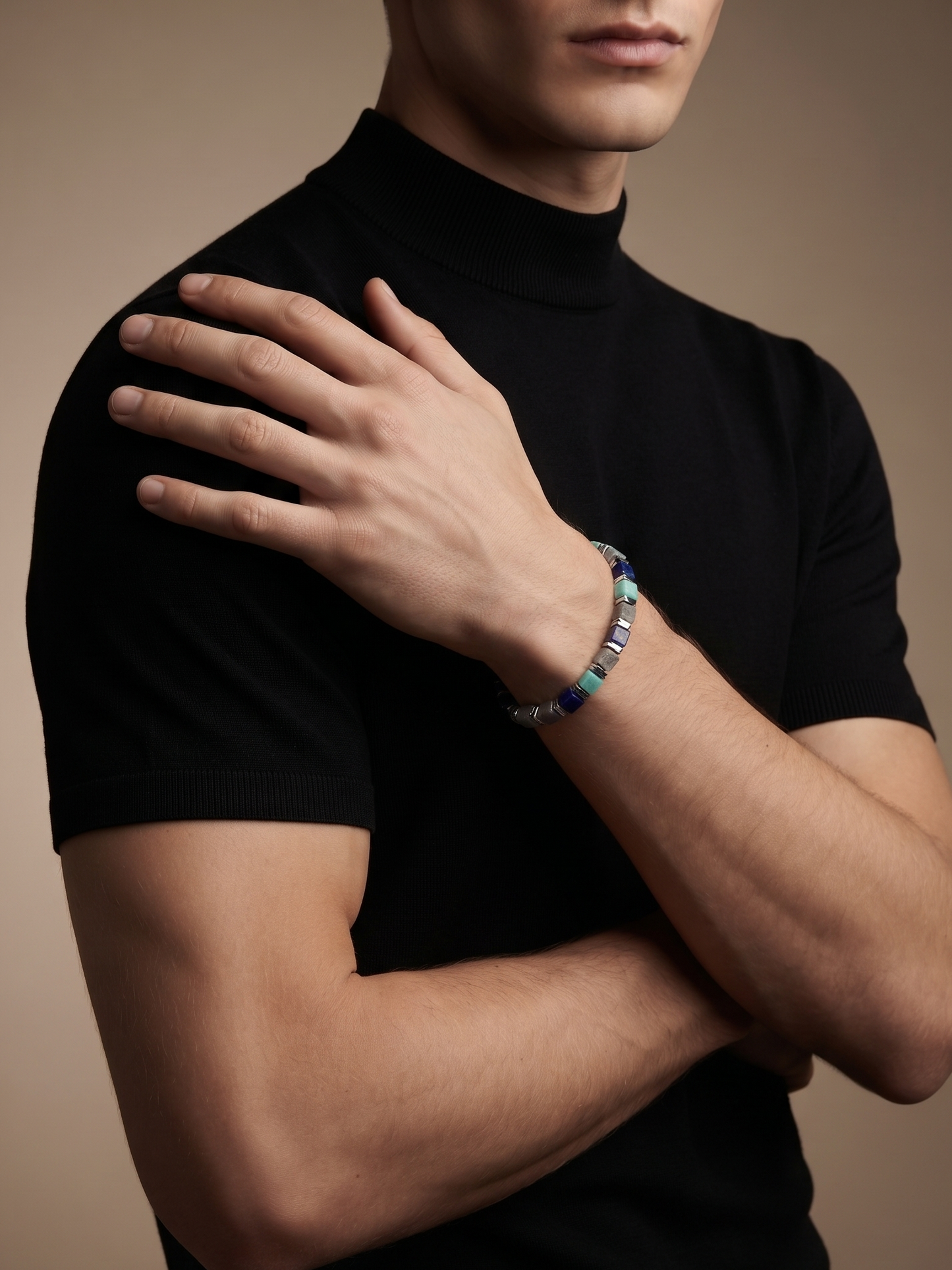 A close-up of a man wearing a black short-sleeve mock neck t-shirt and a beaded bracelet with blue, white, and silver beads on his left wrist, posed against a solid warm brown background.